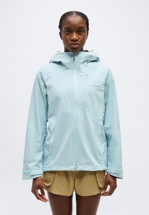 Žena otočená čelom v svetlomodrej kapucňovej pršiplášti a béžových šortkách na jednoduchom bielom pozadí.