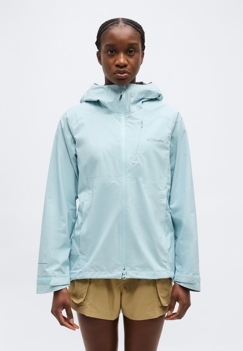 Γυναίκα που κοιτάει μπροστά φορώντας ανοιχτό μπλε αδιάβροχο με κουκούλα και κρεμ σορτς, με φόντο απλό λευκό φόντο.