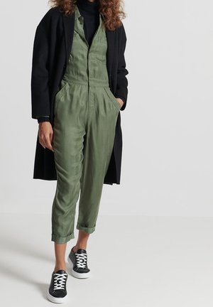 Person trägt olivgrünen Overall mit hochgekrempelten Bündchen, schwarzen langem Mantel, schwarzem Rollkragenpullover und schwarzen Sneakers mit weißen Schnürsenkeln vor einfachem Hintergrund.