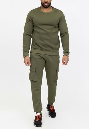 Homme portant un sweat-shirt vert olive et un pantalon cargo avec des baskets noires aux lacets orange, marchant vers l'avant sur un fond blanc.