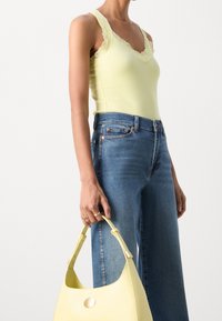 Kvinna som bär en ljusgul linne med spetskant och blå jeans, håller en matchande gul handväska med gulddetaljer.