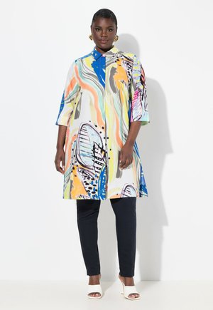 Camicia lunga colorata con motivi astratti in blu, giallo e rosa, con colletto e chiusura a bottoni, abbinata a pantaloni scuri e scarpe bianche.