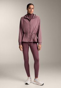 Lichtgewicht, winddicht jack in mauve met een capuchon en een geregen taille; gecombineerd met bijpassende legging en witte sportschoenen.