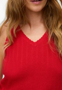 Rød strikket tanktop med en bølget V-hals. Har lodrette striber og et tekstureret mønster. Blødt stof med et let udseende.