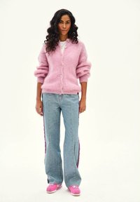 Žena s dlhými kučeravými vlasmi, oblečená v ružovom chlpatom kardigáne, svetlomodrých džínsoch s bočnými pruhmi a ružových teniskách, stojaca na jednoduchom pozadí.