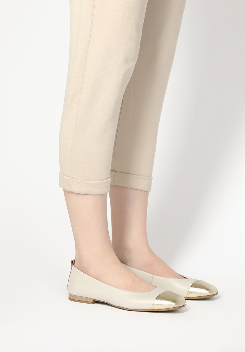 Flache Schuhe mit einem Körper aus cremefarbenem Leder, metallisch goldenem Kappenzunge und einem kleinen Holzabsatz. Kombiniert mit hellen, cropped Hosen.
