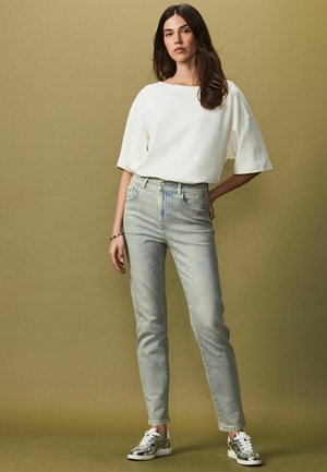 Bílý oversized krátký rukáv top v kombinaci se světlými modrými kalhotami s vysokým pasem a stříbrnými teniskami, umístěný na jemném zeleném pozadí.