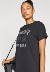 Μαύρο βαμβακερό t-shirt με κοντά μανίκια· διαθέτει γκρι γραφικό κείμενο "1989 NEW YORK" και ραφές με λεπτομέρειες. Φορεμένο με τσάντα με λουρί.