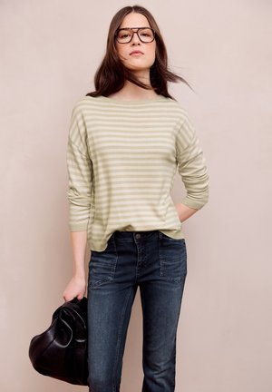 Kvinde med langt brunt hår iført briller, beige stribet sweater, mørke jeans, der holder en sort lædertaske mod beige baggrund.