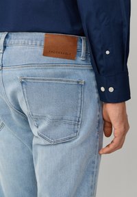 Man draagt lichtblauwe jeans met een bruine Façonnable leren label en een donkerblauw shirt, staat met de rechterhand zichtbaar naast de heup.