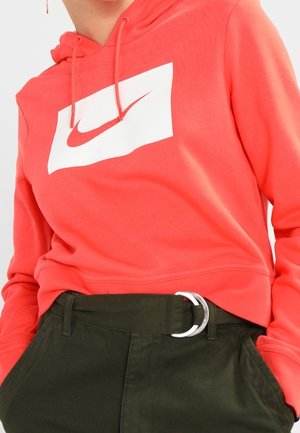Personne portant un sweat à capuche Nike rouge corail avec un logo blanc et un pantalon vert foncé avec une boucle de ceinture en D argentée, les mains dans les poches.