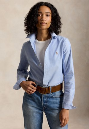 Γυναίκα με μαύρα σγουρά μαλλιά φοράει ένα γαλάζιο πουκάμισο με κουμπιά πάνω από ένα λευκό μπλουζάκι, μπλε τζιν και καφέ ζώνη, στέκεται μπροστά σε ουδέτερο φόντο.