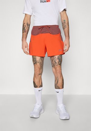 Homme portant un short de course orange et marron, des chaussettes Nike blanches et des baskets blanches, se tenant contre un fond uni.