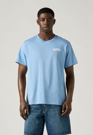 Hombre negro de pie mirando hacia adelante, vistiendo una camiseta Levi's azul claro y pantalones cortos vaqueros azules sobre un fondo liso.