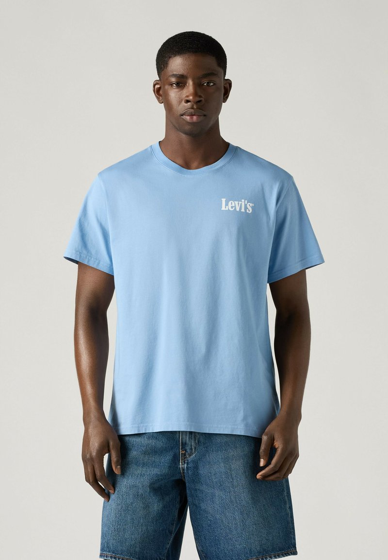 Ein schwarzer Mann steht frontal, trägt ein hellblaues Levi's T-Shirt und blaue Jeansshorts vor einem einfarbigen Hintergrund.