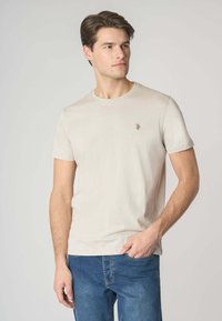 T-shirt in cotone beige chiaro con collo rotondo, maniche corte e un piccolo logo ricamato sul petto. Abbinato a jeans in denim blu.