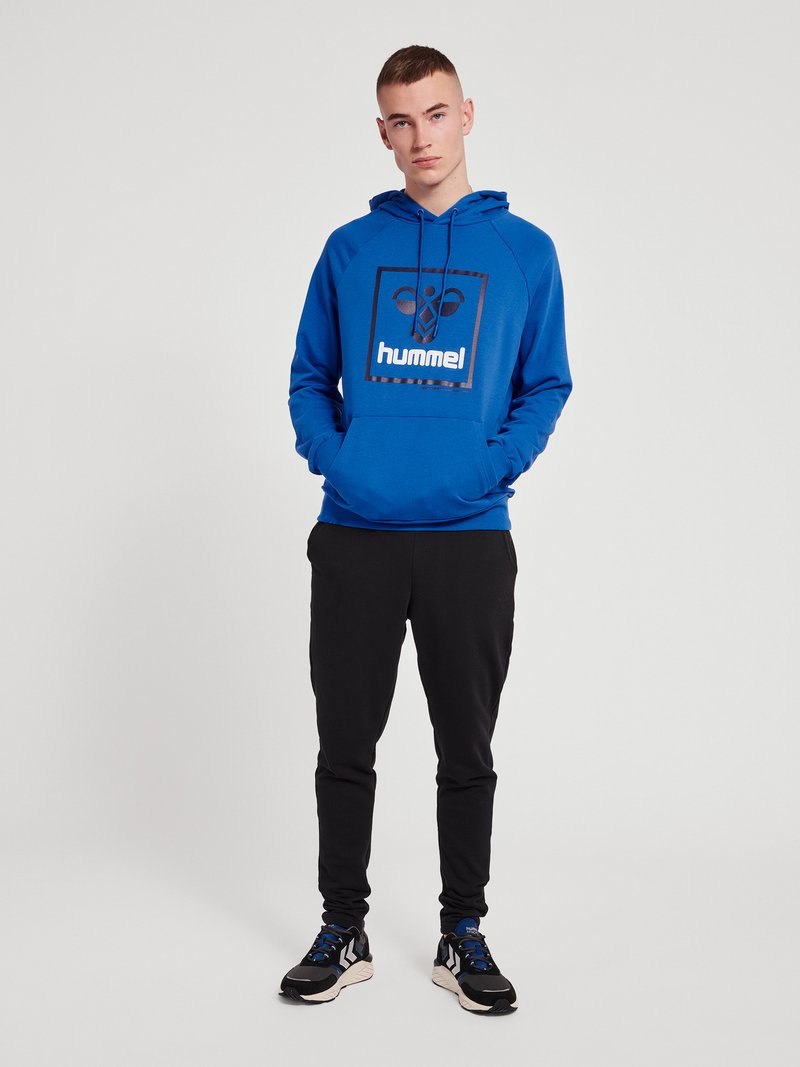 Jonge man staat met handen in de zakken, draagt een blauwe hummel hoodie, zwarte broek en zwart-blauwe sneakers tegen een effen achtergrond.