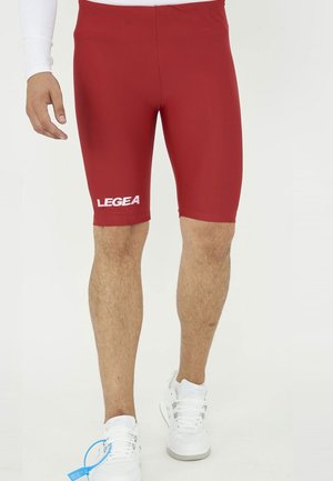 Gambe maschili indossano pantaloncini sportivi rossi fino al ginocchio con il logo "LEGEA" sulla coscia sinistra e scarpe da ginnastica bianche su sfondo semplice.
