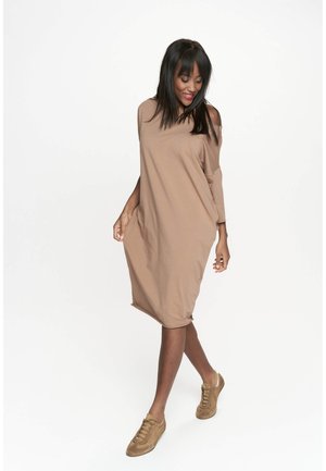 Brą dress w kolorze brązowym, oversize, z odsłoniętymi ramionami, długimi rękawami, miękką teksturą i luźnym krojem, w zestawieniu z jasnobrązowymi butami.