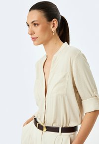 Beżowa koszula z guzikiem, z podwiniętymi rękawami, dwiema kieszeniami na piersi i luźnym krojem, zestawiona z brązowym paskiem z złotą klamrą.
