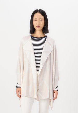 Châle léger, ouvert sur le devant, en beige doux avec un motif circulaire. Le tissu texturé tombe librement sur un haut rayé et un pantalon blanc.