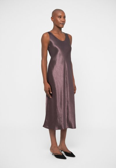 Ärmelloses, knielanges Kleid aus glänzendem, dunkellila Stoff mit glatter Textur. Model trägt schwarze Slipper und steht vor einem neutralen Hintergrund.