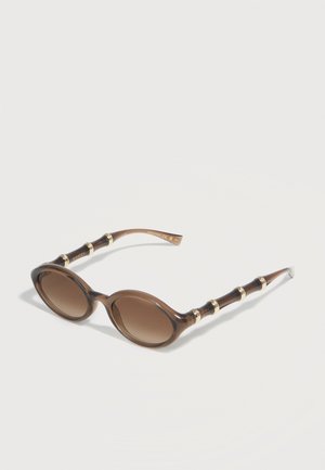 Lunettes de soleil ovales brunes avec verres dégradés et branches texturées inspirées du bambou, ornées d'accents dorés, présentées sur un fond blanc.