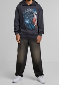 Zwarte hoodie met een grafisch ontwerp van een stekelhart, ruime donkere jeans en witte sneakers. De hoodie heeft een voorzak en geribde mouwen.
