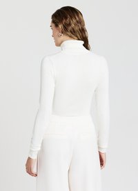 Langärmliger weißer Rollkragenpullover mit taillierter Passform, Rippstruktur und weichem Material. Kombiniert mit hoch taillierten weißen Stoffhosen.