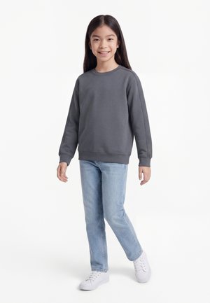 Jeune fille souriante, portant un sweat-shirt gris foncé, un jean bleu clair et des baskets blanches, debout devant un fond blanc.