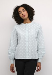 Lichtblauwe blouse met textuur stippenpatronen, een ronde kraag, knoopsluiting en kanten accenten op de mouwen. Elastische manchetten.