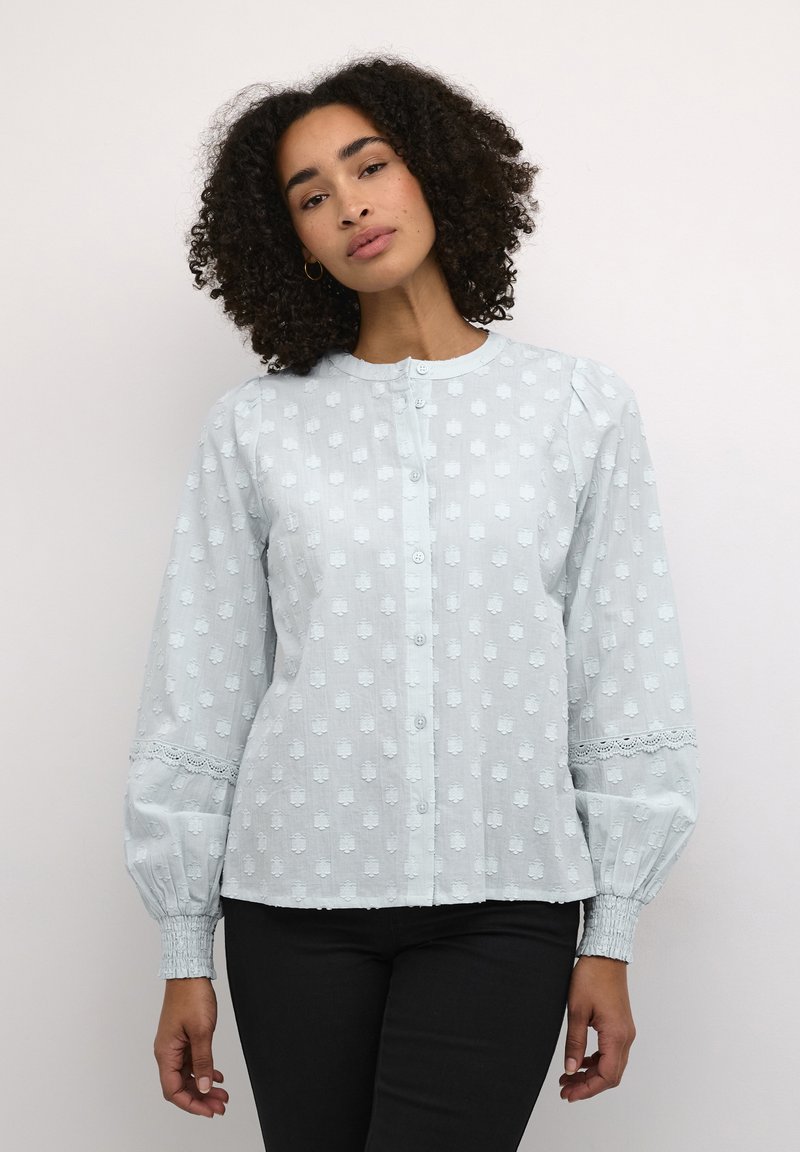 Lichtblauwe blouse met textuur stippenpatronen, een ronde kraag, knoopsluiting en kanten accenten op de mouwen. Elastische manchetten.