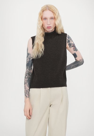 Dark brown sleeveless turtleneck top in a soft knit fabric, paired with light beige wide-leg trousers, showing intricate arm tattoos.