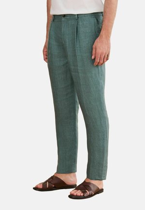Mann trägt grüne Leinenhose und braune Ledersandalen und steht mit entspannten Händen an den Seiten vor einem schlichten Hintergrund.