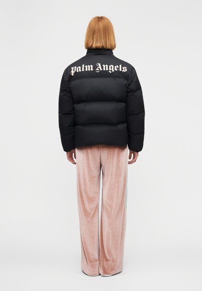Μαύρο μπουφάν puffer με την επιγραφή "Palm Angels" στην πλάτη σε μεγάλα λευκά γράμματα. Φορεμένο πάνω από ροζ βελούδινο παντελόνι με λεία υφή.
