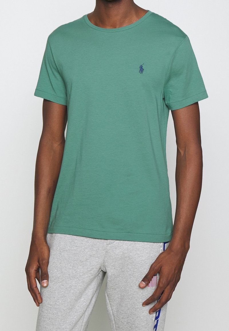 Man som bär en grön T-shirt med kort ärm och en liten blå logotyp på bröstet, samt ljusgråa sweatpants med blå och röd text på benen.