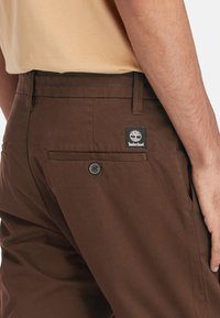 Pantaloni marroni in cotone con una texture liscia. Presentano una tasca posteriore con bottone e un'etichetta con il logo Timberland sulla vita.