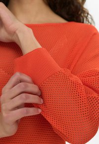 Atvērtas adīšanas oranžs džemperis ar ribotām aprocēm un kaklu. Audums ir elpojošs, ar brīvu dizainu un režģa rakstu.