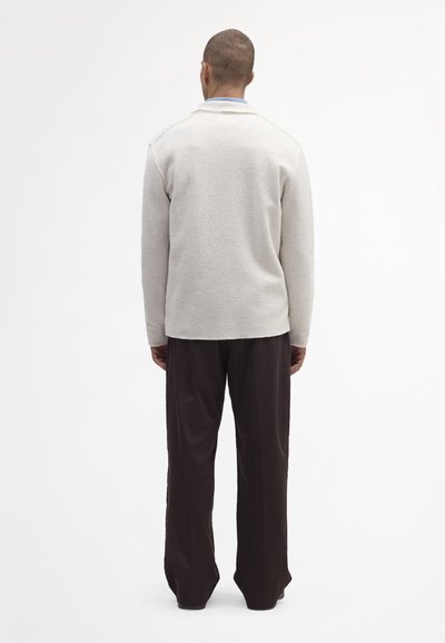 Hellgraue Fleece-Jacke mit umgeschlagenem Kragen, langen Ärmeln und lässiger Passform, kombiniert mit dunkelbraunen weit geschnittenen Hosen.