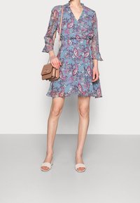 Sininen kukkakuosiset mekko, jossa on pitkät, levenevät hihat; vyötärö on korostettu; parina pieni ruskea laukku ja vaaleat liukumekanavat.