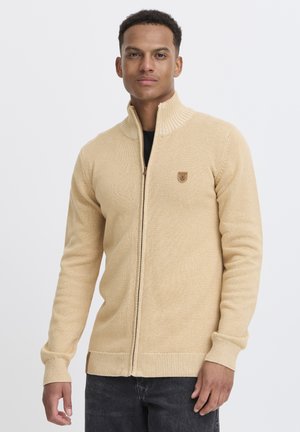 Beige zip-up genser med høy krage, ribbede ermer og et lite emblem på brystet. Mykt strikket stoff med en lett glitrende tekstur.
