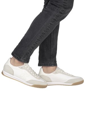 Personne portant un jean noir ajusté et des baskets blanches avec des accents en daim beige, debout avec un pied croisé sur l'autre.