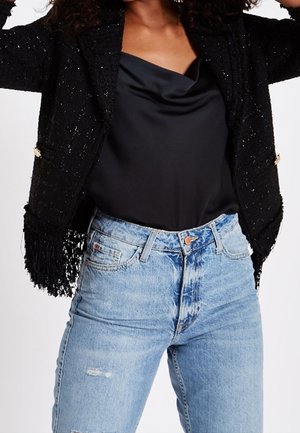 Schwarze, strukturierte Fransenjacke über einem schwarzen Satinoberteil, kombiniert mit hoch taillierten, hellblauen, verwaschenen Jeans an einer stehenden Person.