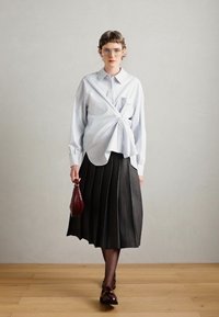 Chemise bleu clair avec un détail de nœud à l'avant, jupe plissée noire, chaussures marron et sac à main bordeaux ; le modèle se tient sur un sol en bois devant un mur uni.