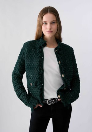 Femme aux longs cheveux raides portant une veste texturée vert foncé sur une chemise blanche et un pantalon noir avec une ceinture cloutée.