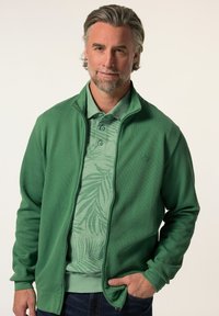 Giacca verde con zip presenta una texture a coste e un colletto alto, indossata sopra un polo verde chiaro con un motivo di foglie tropicali.