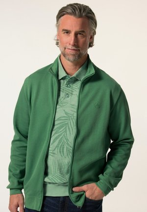 Green zip-up jacket features ribbed texture and a high collar, layered over a light green polo with a tropical leaf pattern.