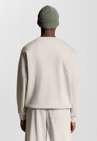 Uomo di spalle che indossa un completo felpa e pantaloni beige chiaro con un berretto lavorato a maglia verde oliva su uno sfondo neutro.