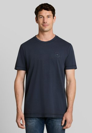 Man draagt een effen donkerblauw T-shirt met korte mouwen en blauwe spijkerbroek, staand tegen een neutrale grijze achtergrond.