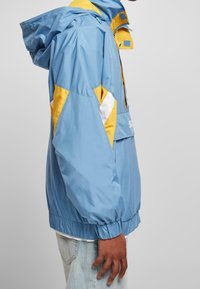 Giacca azzurro chiaro con accenti gialli e bianchi, dotata di cappuccio, polsini elasticizzati e chiusura con zip frontale. Il materiale sembra liscio e leggero.